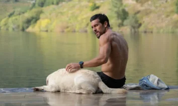 Mario Casas em cena de Agente Zeta, suspense do Prime Video, sentado à beira de um lago ao lado de um cachorro