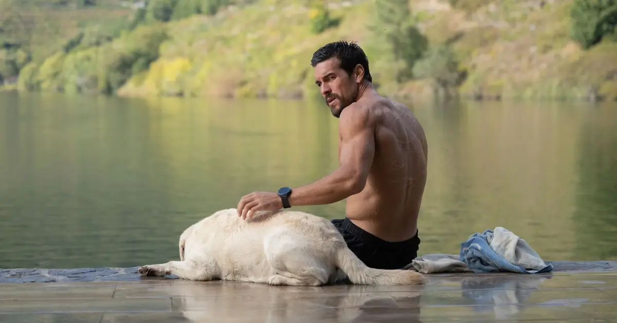 Mario Casas em cena de Agente Zeta, suspense do Prime Video, sentado à beira de um lago ao lado de um cachorro