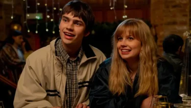 Spike Fearn e Angourie Rice sorriem e olham para cima em uma cena do filme de comédia romântica "Procurando Emily". Spike usa uma jaqueta bege sobre camisa xadrez e Angourie usa uma jaqueta escura. O fundo é um ambiente interno aconchegante com luzes decorativas.