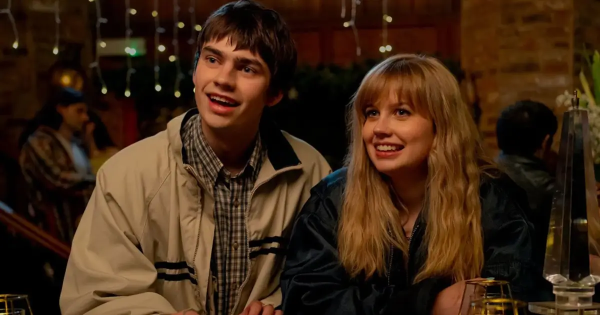 Spike Fearn e Angourie Rice sorriem e olham para cima em uma cena do filme de comédia romântica "Procurando Emily". Spike usa uma jaqueta bege sobre camisa xadrez e Angourie usa uma jaqueta escura. O fundo é um ambiente interno aconchegante com luzes decorativas.