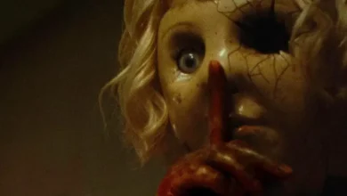 Cena do trailer de Dolly – A Boneca Maldita (2026), mostrando a boneca com máscara rachada fazendo sinal de silêncio.