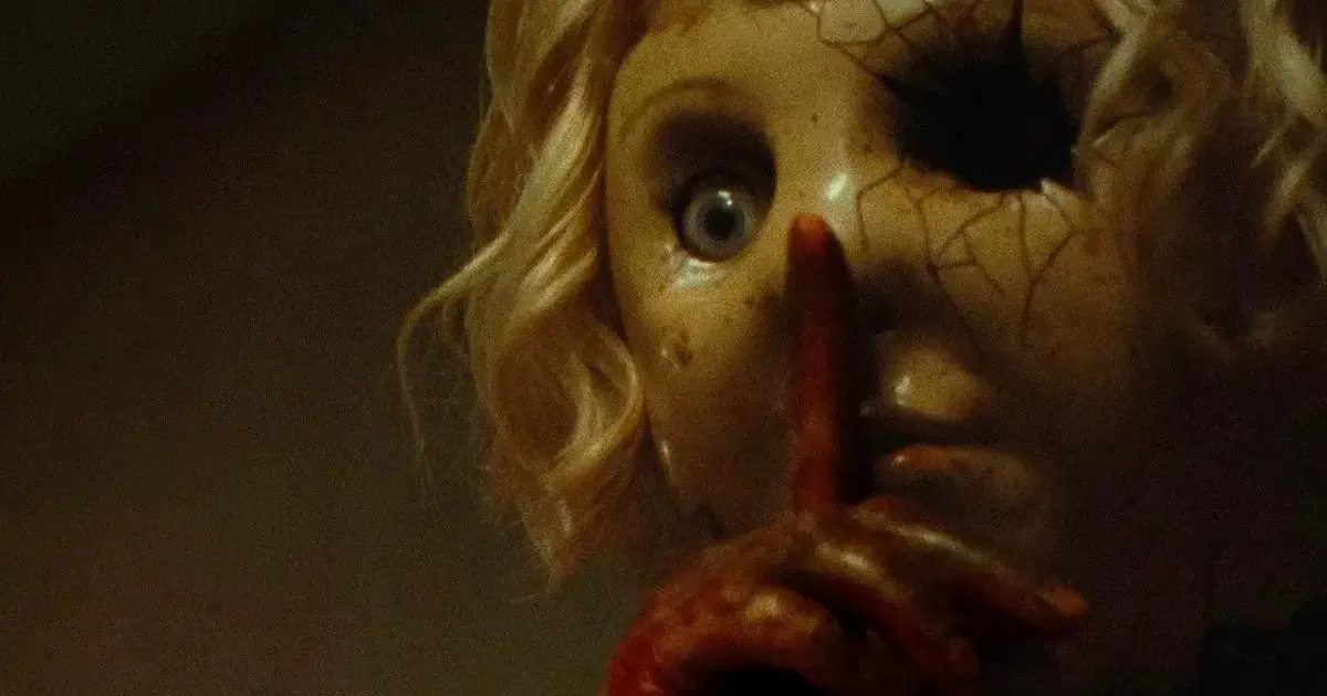 Cena do trailer de Dolly – A Boneca Maldita (2026), mostrando a boneca com máscara rachada fazendo sinal de silêncio.