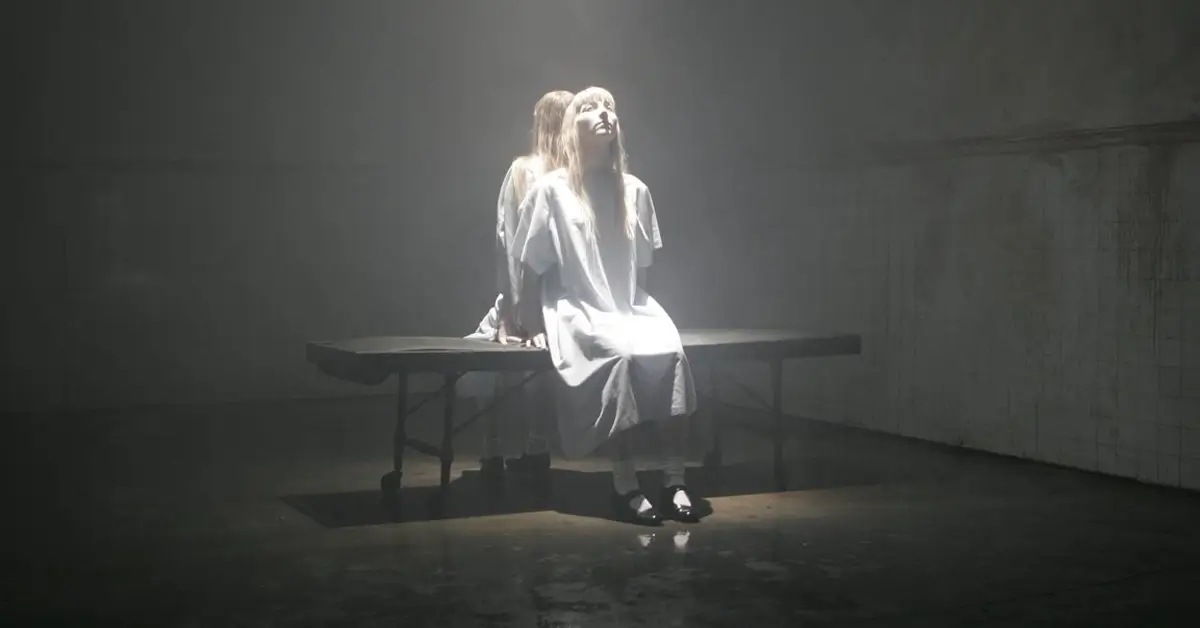 Cena sombria do trailer de Phantasmagoria The Visions of Lewis Carroll com mulher em ambiente hospitalar iluminado, estilo perturbador de Marilyn Manson