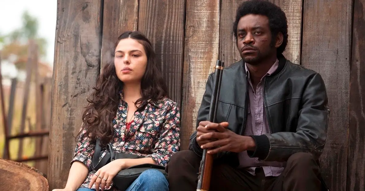 Fabrício Boliveira como João de Santo Cristo e Isis Valverde como Maria Lúcia em cena do filme Faroeste Caboclo (2013), sentados em frente a uma construção de madeira.