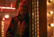 Hunter Schafer como Cora em Blade Runner 2099 (2026), em cenário futurista iluminado por luzes vermelhas e amarelas.