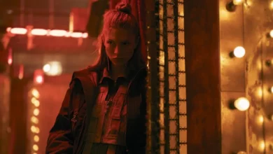 Hunter Schafer como Cora em Blade Runner 2099 (2026), em cenário futurista iluminado por luzes vermelhas e amarelas.