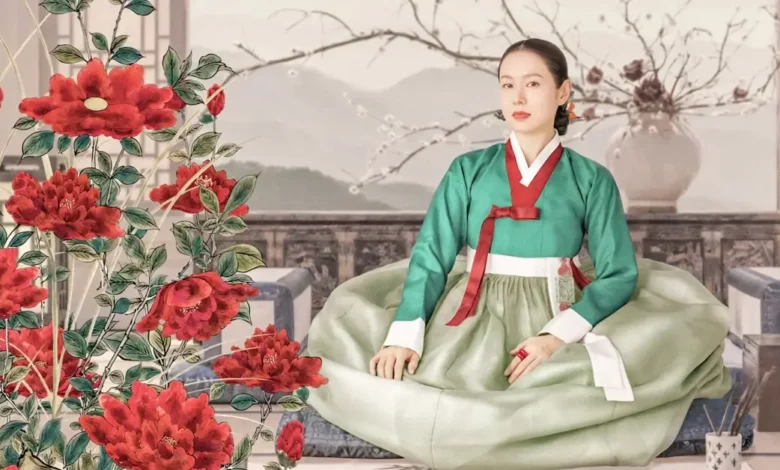 Son Ye-jin interpreta Lady Cho em Escândalo e Intriga (The Scandal), nova minissérie coreana da Netflix ambientada na era Joseon; a atriz aparece sentada em um cenário tradicional cercado por flores vermelhas.