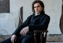Kit Harington como Sydney Carton sentado em uma cadeira de madeira, usando traje preto de época, na minissérie Um Conto de Duas Cidades (A Tale of Two Cities), do MGM+