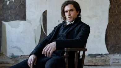 Kit Harington como Sydney Carton sentado em uma cadeira de madeira, usando traje preto de época, na minissérie Um Conto de Duas Cidades (A Tale of Two Cities), do MGM+