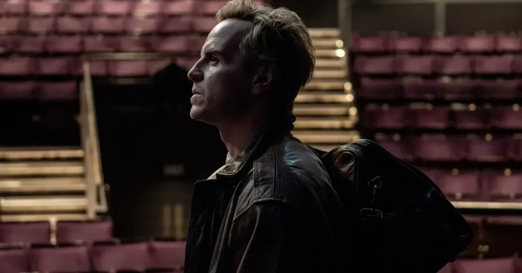 Andrew Scott em Elsinore (2027), sentado em um teatro vazio com uma bolsa de viagem sobre o ombro.