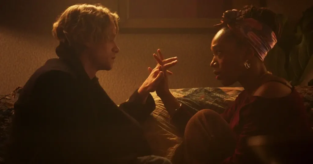 Tom Rhys Harries e Naomi Ackie em cena dramática de Cara-de-Barro (2026), conversando frente a frente em um quarto com iluminação quente.