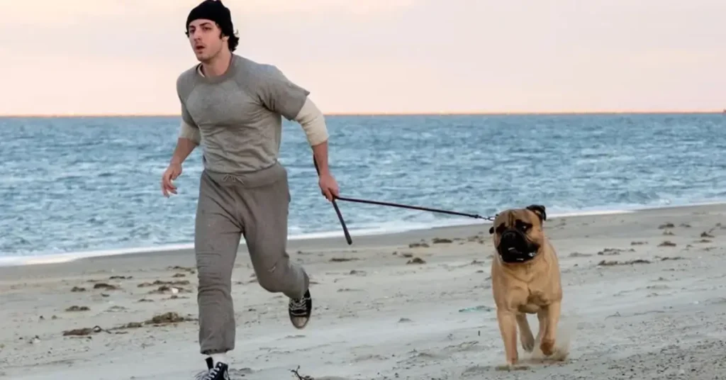 Anthony Ippolito como Sylvester Stallone correndo na praia com o cachorro Butkus em cena de Eu Sou o Rocky (I Play Rocky, 2026)